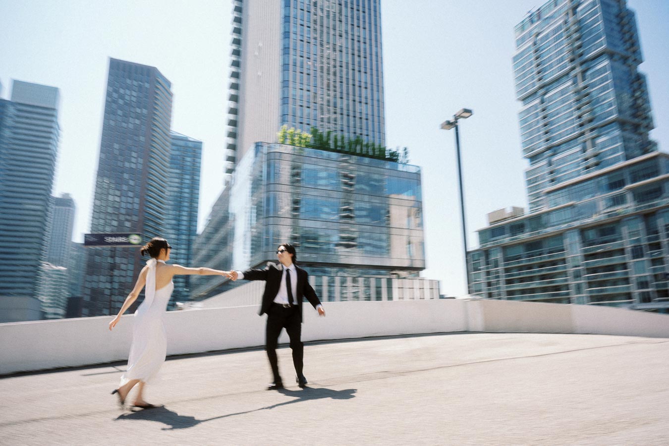 A couple in formal outfits holding hands and walking on a rooftop with tall glass buildings in downtown Toronto.