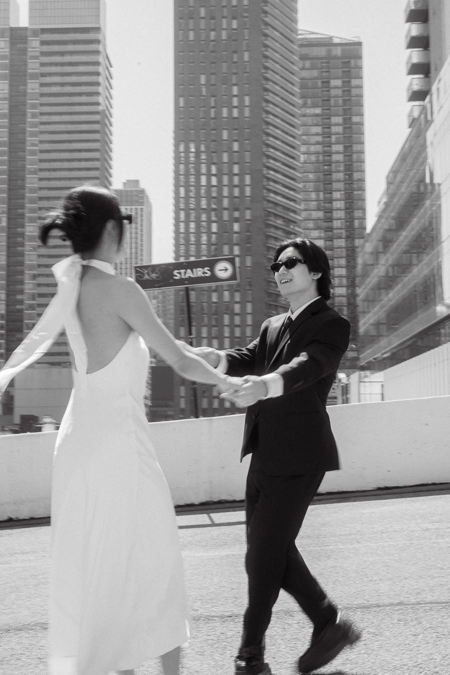 Couple spinning and laughing together on a rooftop with tall city buildings in the background, both wearing sunglasses and formal outfits.