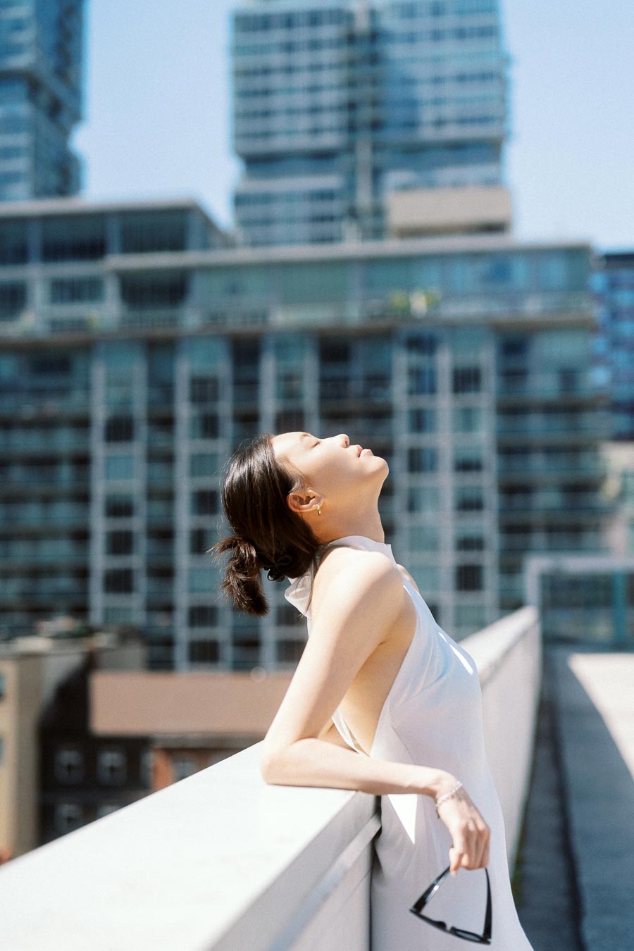 Woman in white dress leaning on rooftop railing, eyes closed, with city buildings in the background.
