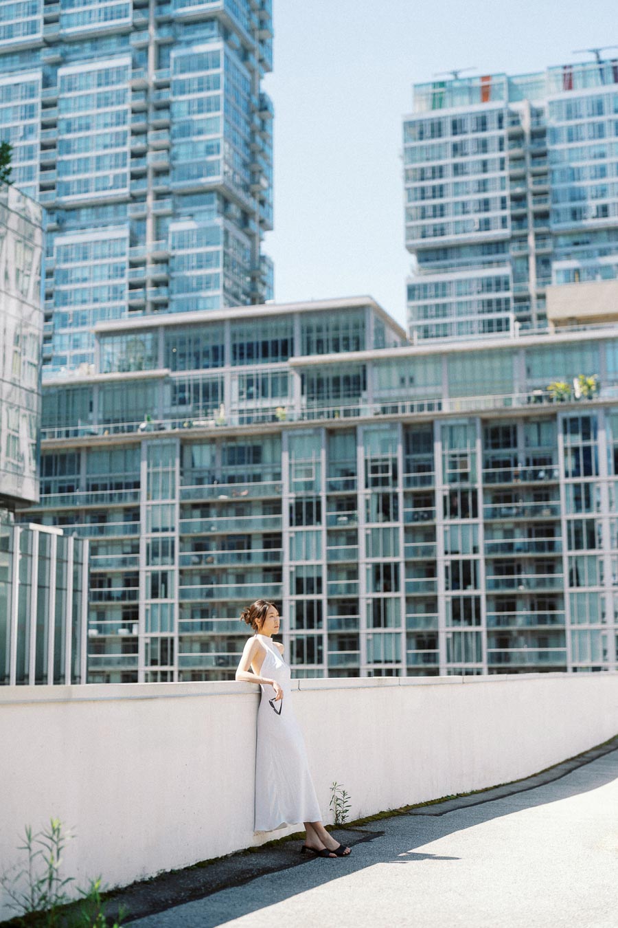 Woman in white dress leaning on rooftop wall with modern glass buildings in background on a sunny day.