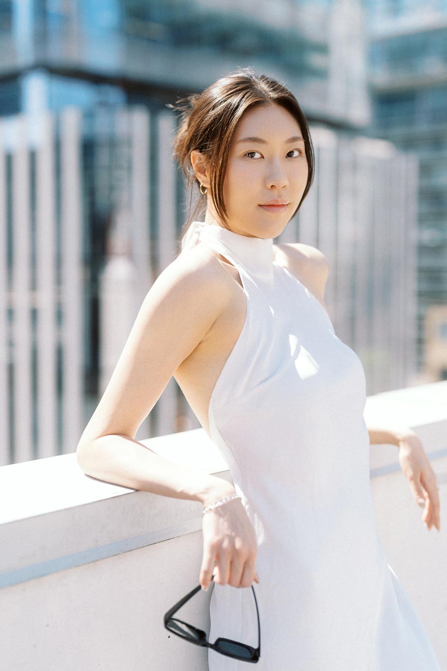 Woman in white dress holding sunglasses on a rooftop with glass skyscrapers behind her in downtown Toronto.