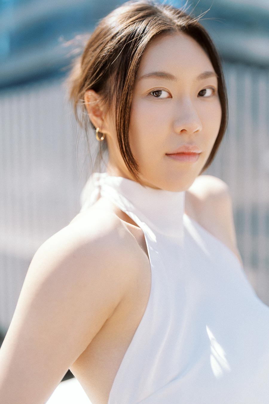 Woman in a white halter dress posing outdoors with soft sunlight and city buildings behind her.