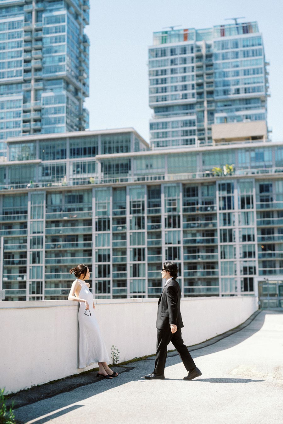 Couple on a rooftop, one leaning on a wall in a white dress, the other walking toward them in a black suit, with tall glass buildings in the background.
