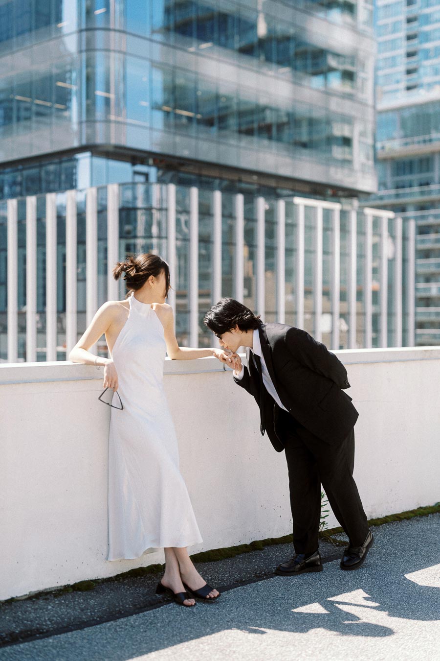 Couple on a rooftop, one in a white dress and one in a black suit, sharing a hand kiss against a backdrop of modern glass buildings.
