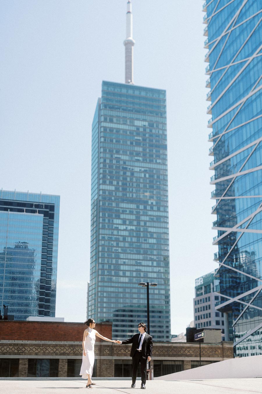 Couple holding hands on a rooftop in downtown Toronto with modern skyscrapers in the background, both dressed in formal attire.
