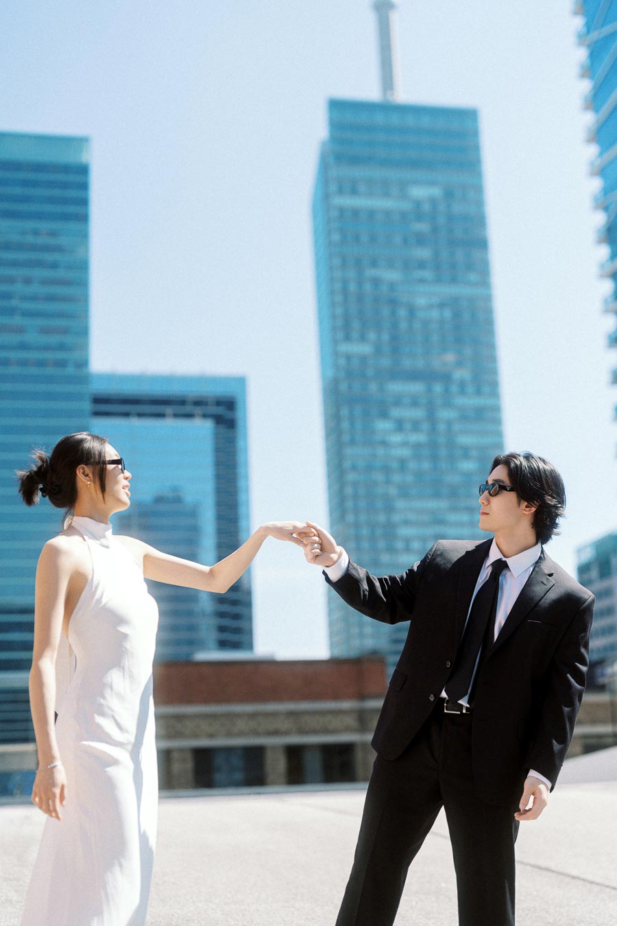 Couple in formal attire holding hands on a rooftop, surrounded by glass skyscrapers under bright daylight in downtown Toronto.