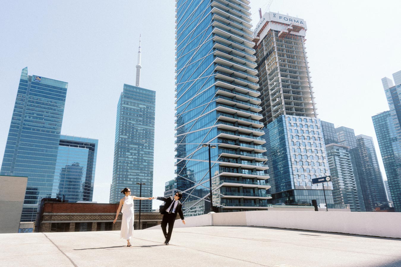 A couple walks holding hands on a rooftop parking garage with Toronto skyscrapers in the background.