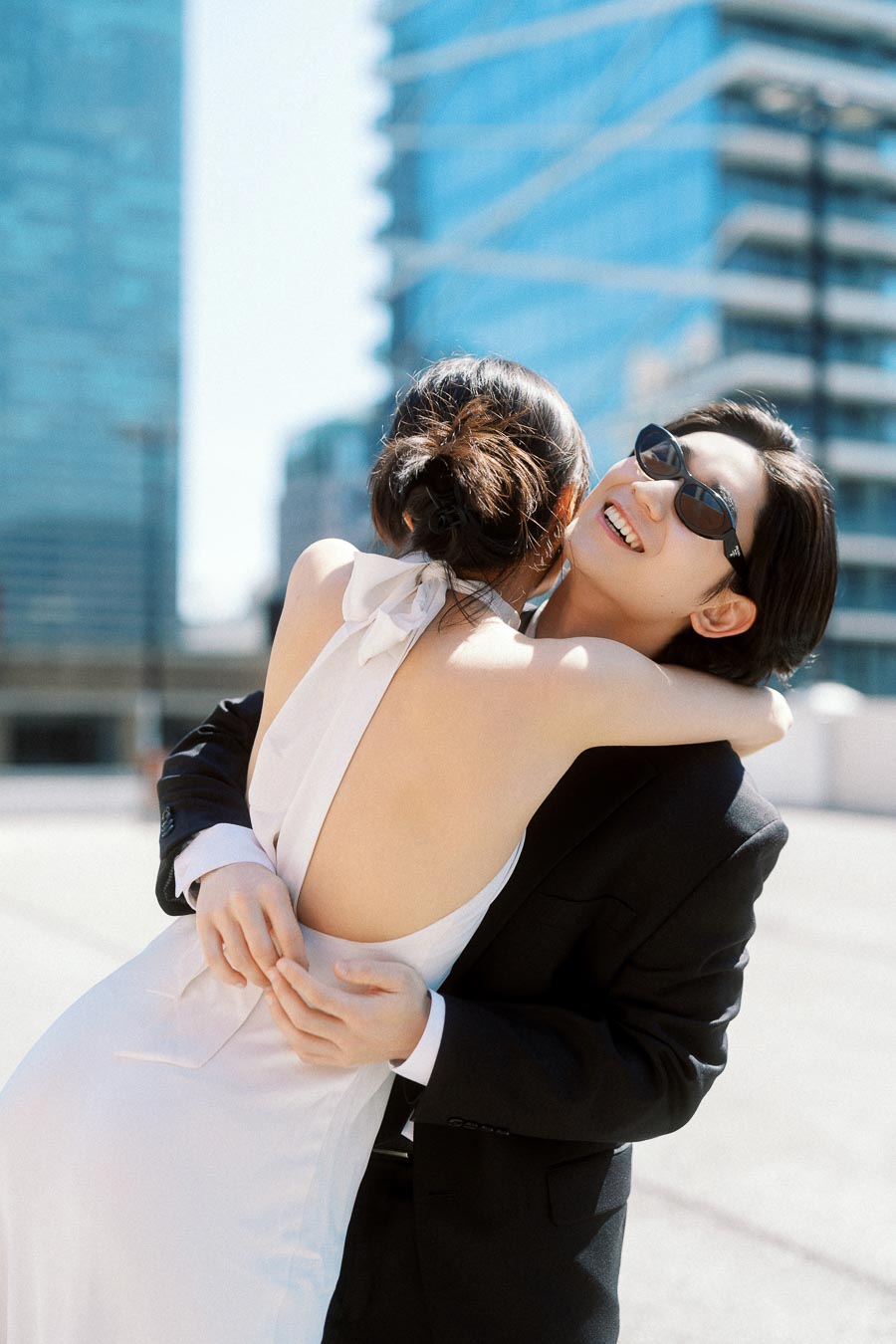 Couple embracing and smiling on a rooftop, one in a white dress and the other in a black suit with sunglasses, city buildings in background.