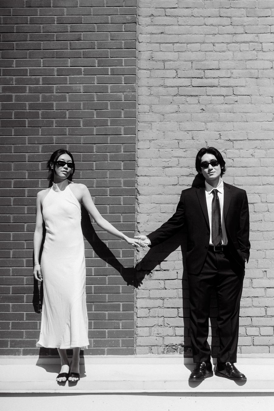 A couple stands against two contrasting brick walls, holding hands and wearing sunglasses, with one in a white dress and one in a black suit.