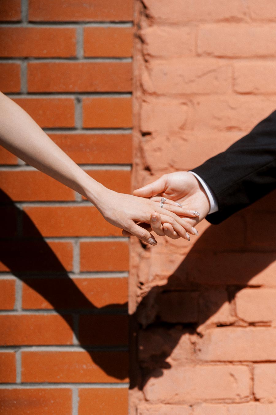 Two people holding hands in sunlight, one wearing an engagement ring, with a red brick wall background.