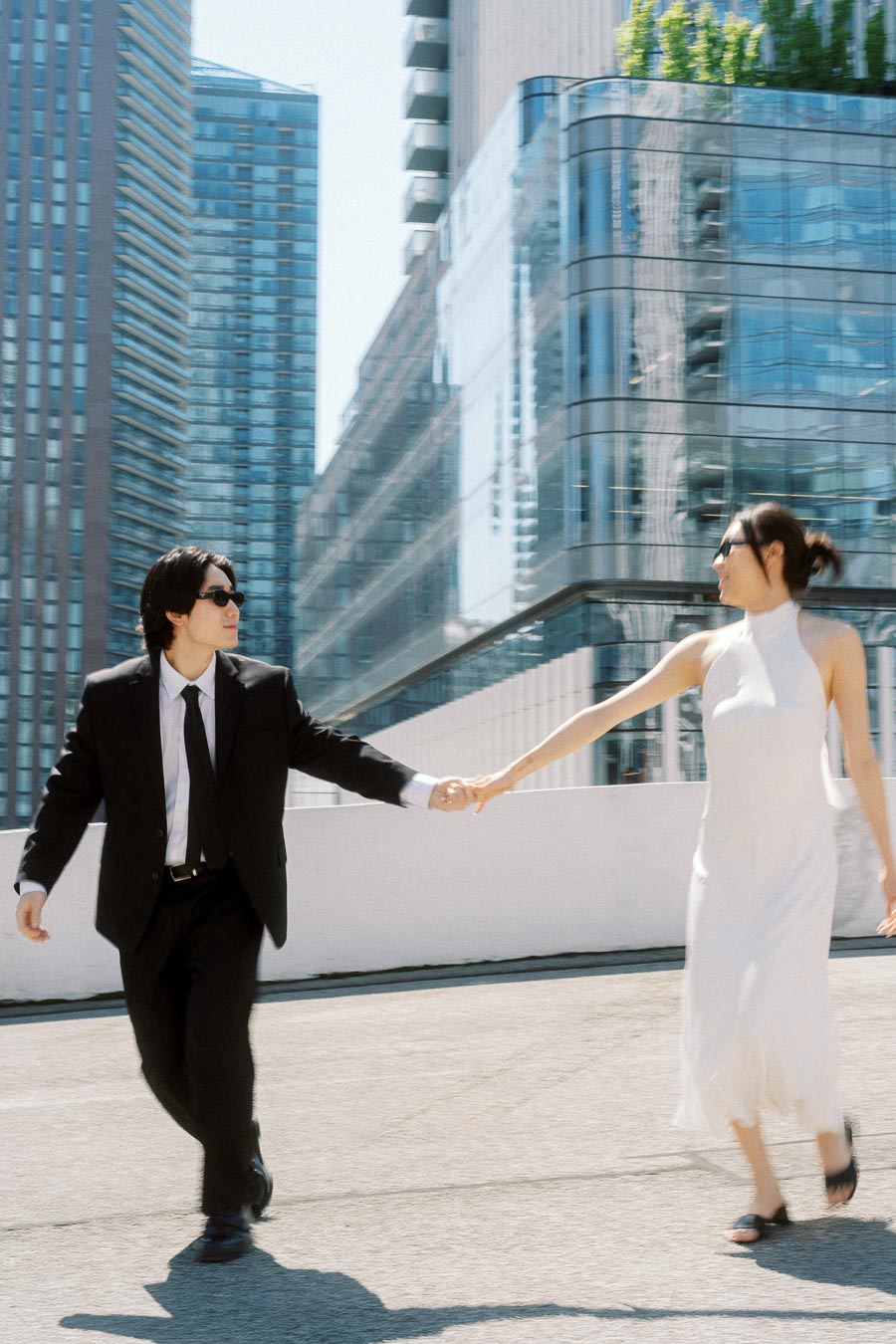 Couple in formal attire holding hands and walking on a rooftop with tall glass buildings in downtown Toronto.