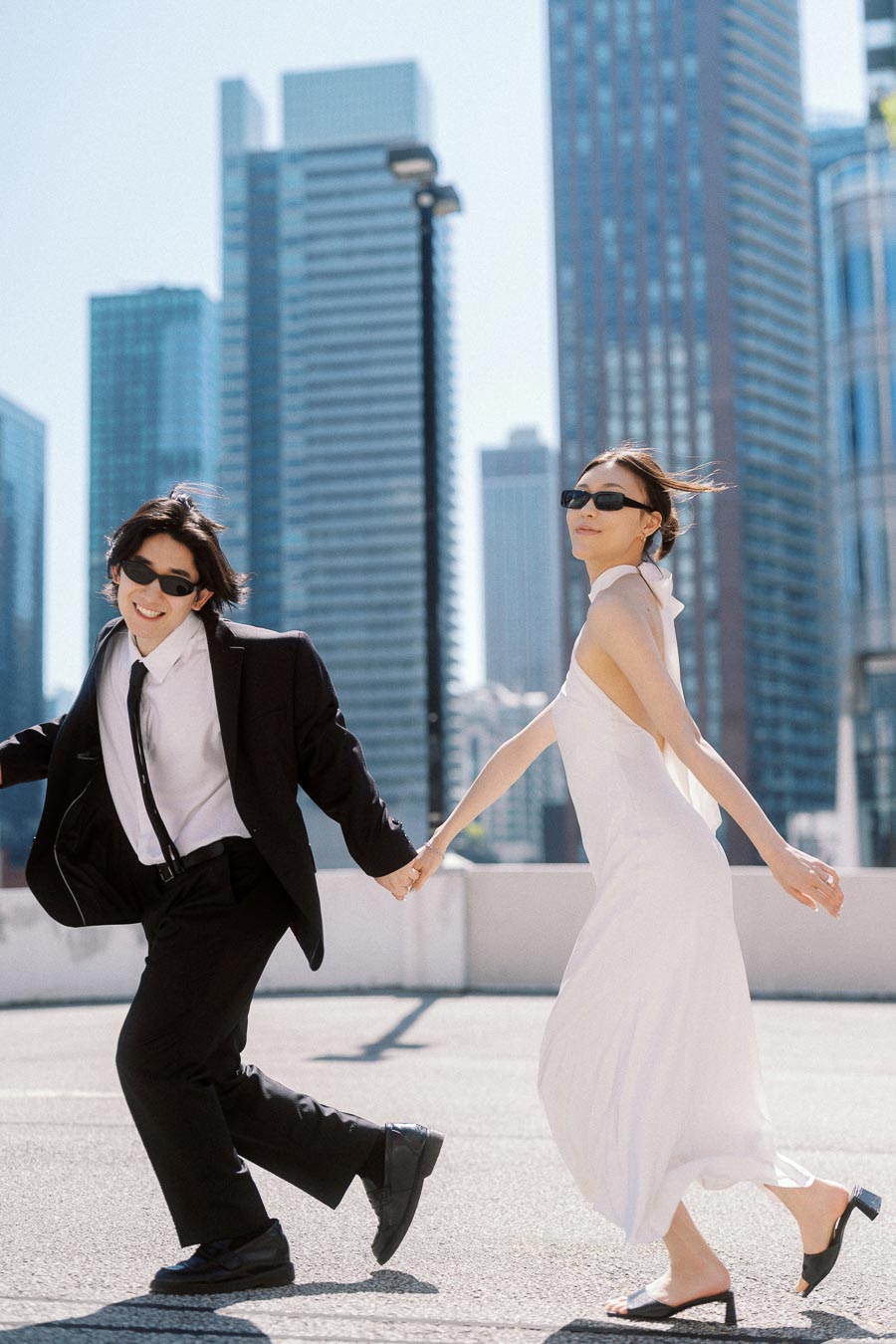 Couple holding hands and running together on a rooftop in downtown Toronto, both wearing sunglasses, with tall city buildings in the background.