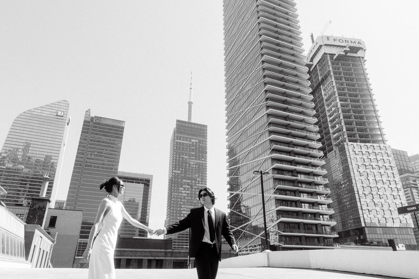 Couple holding hands on a rooftop parking garage, modern Toronto skyscrapers in the background, both wearing sunglasses, one in a white dress, the other in a black suit.