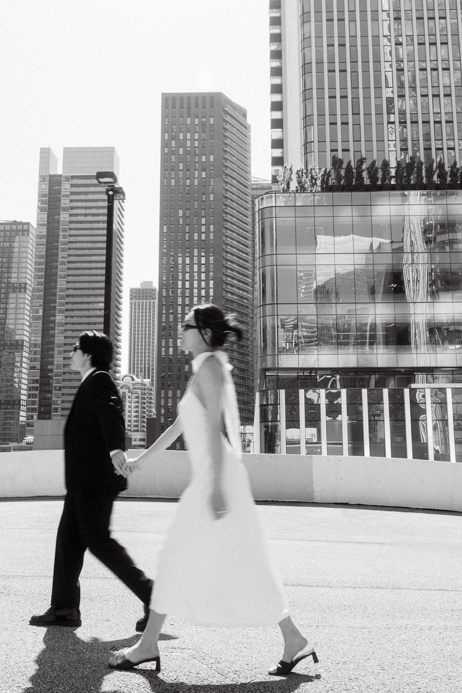 Couple holding hands and walking across a rooftop parking garage, with tall Toronto city buildings in the background, both wearing formal outfits.