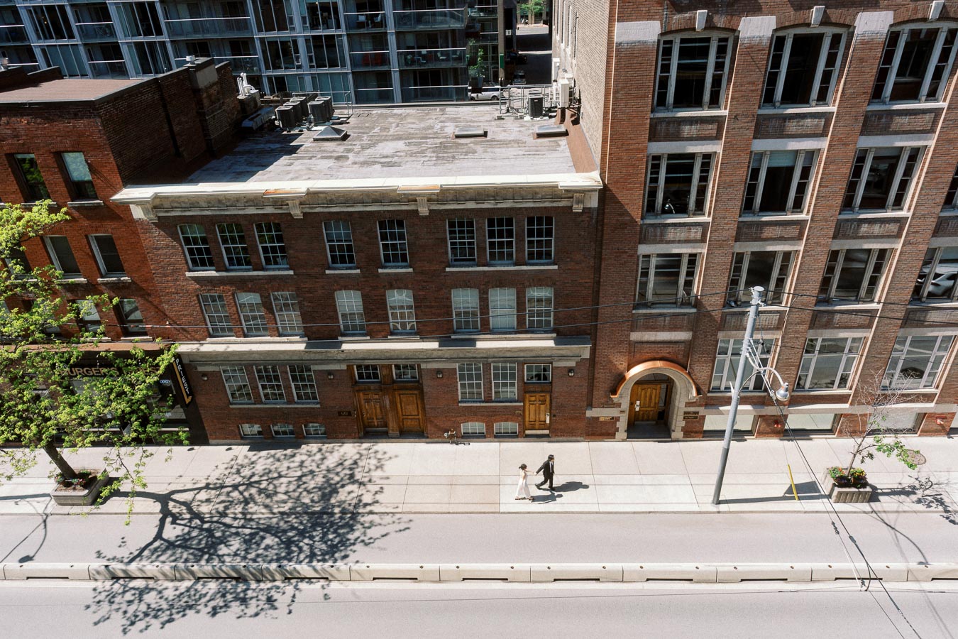 Couple walking hand in hand on a sunny sidewalk in downtown Toronto, surrounded by old brick buildings and city shadows.