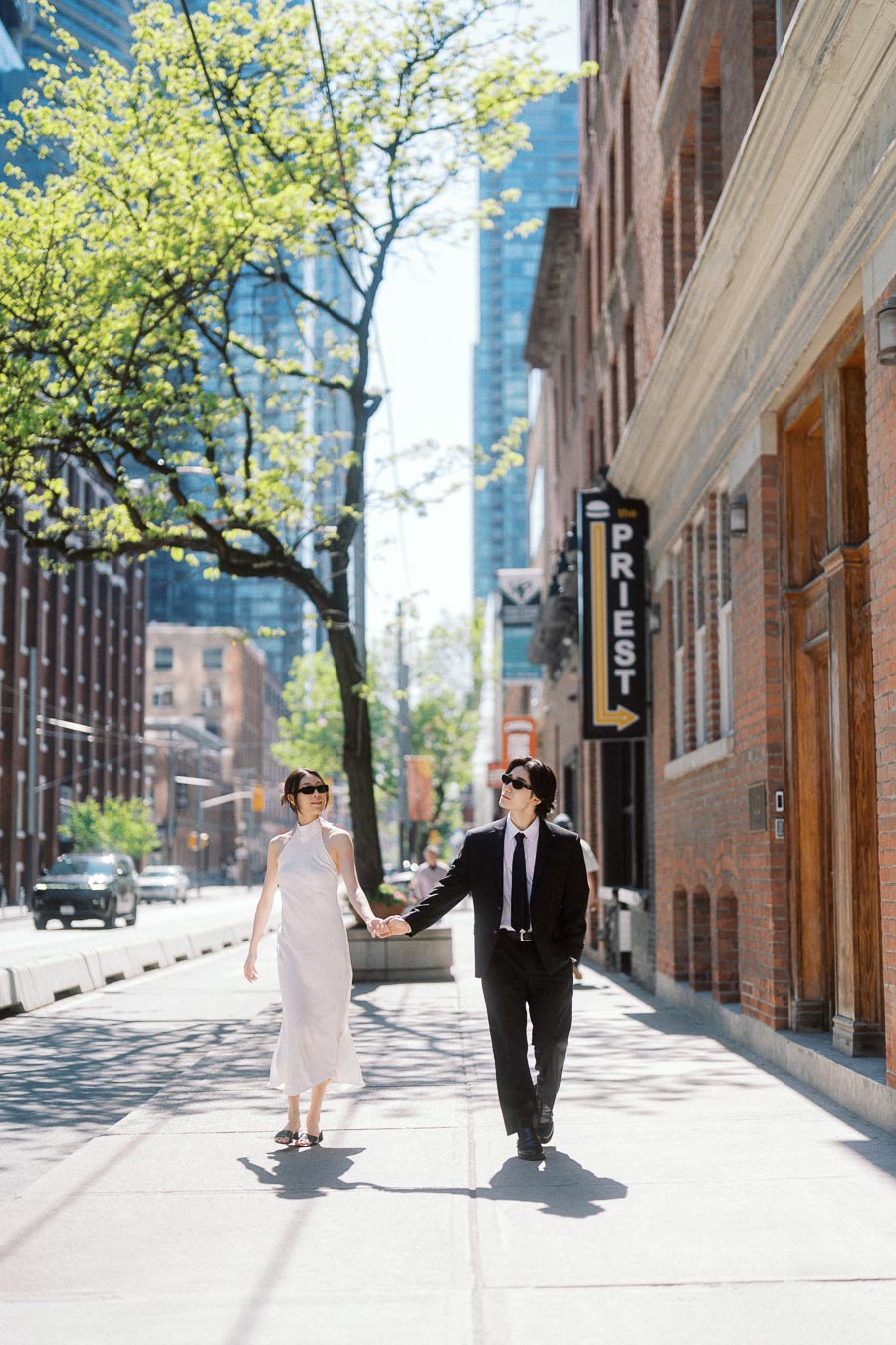 Couple holding hands and walking on a sunny downtown Toronto sidewalk, dressed in a white dress and black suit, with city buildings and trees in the background.