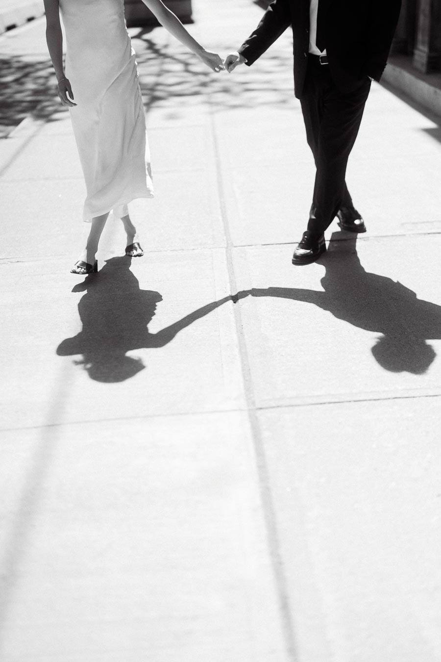 Couple holding hands, walking on a city sidewalk, with their shadows clearly visible on the pavement in dramatic sunlight.