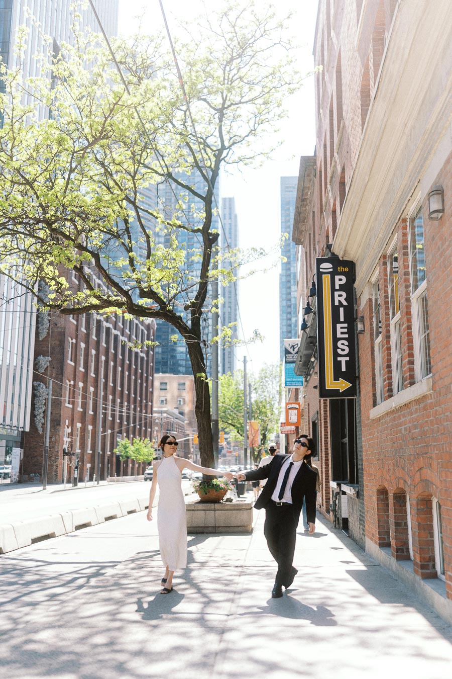 Couple holding hands and walking together on a bright city sidewalk, surrounded by tall buildings and a leafy tree in downtown Toronto.
