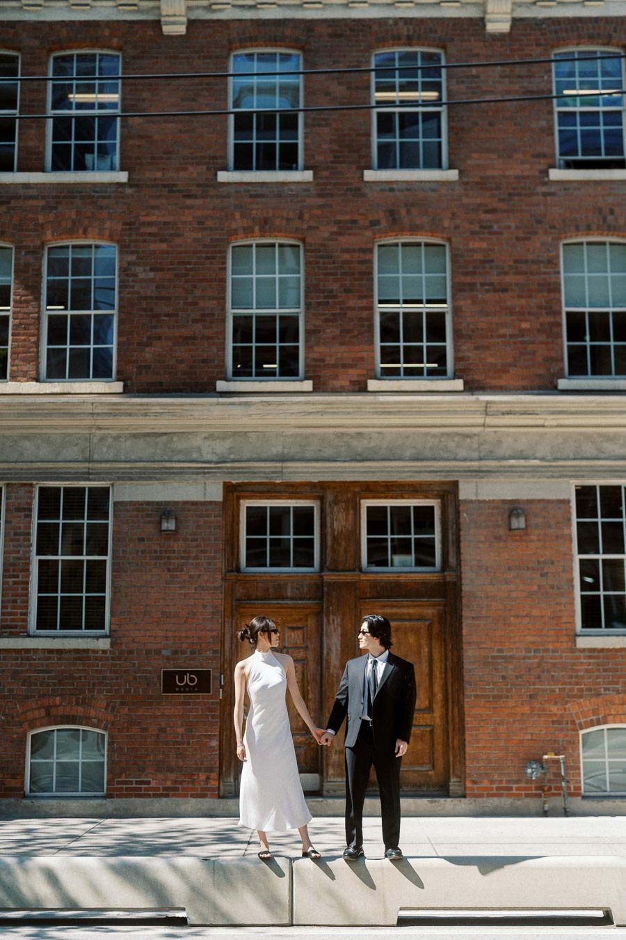 Couple holding hands and looking at each other in front of a large brick building with wooden doors, sunlight casting soft shadows.