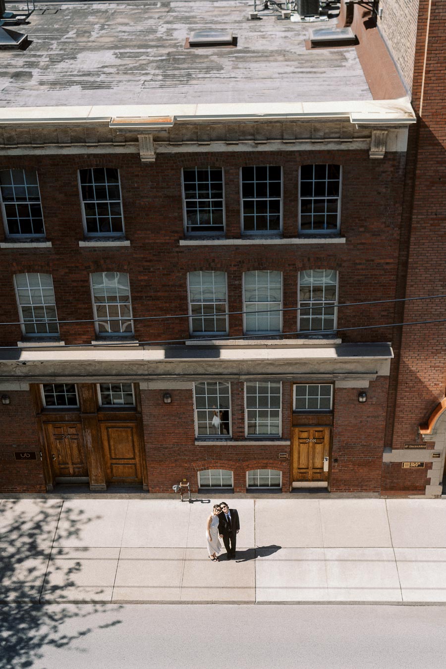 Couple dressed in white and black stands close together on a sunlit sidewalk in front of a red brick building.