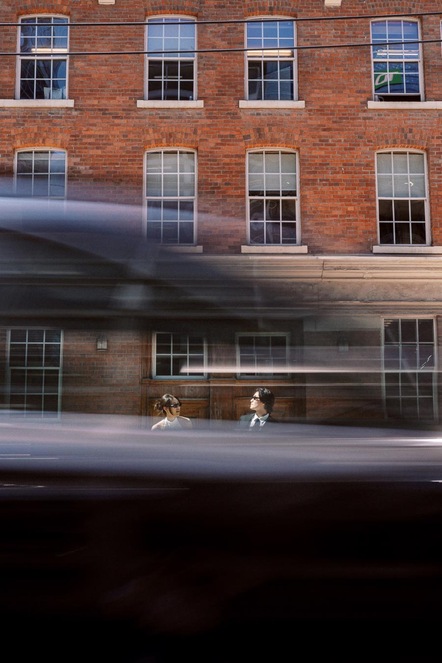 Couple standing in front of a red brick building, partially obscured by blurred motion of a passing vehicle in downtown Toronto.