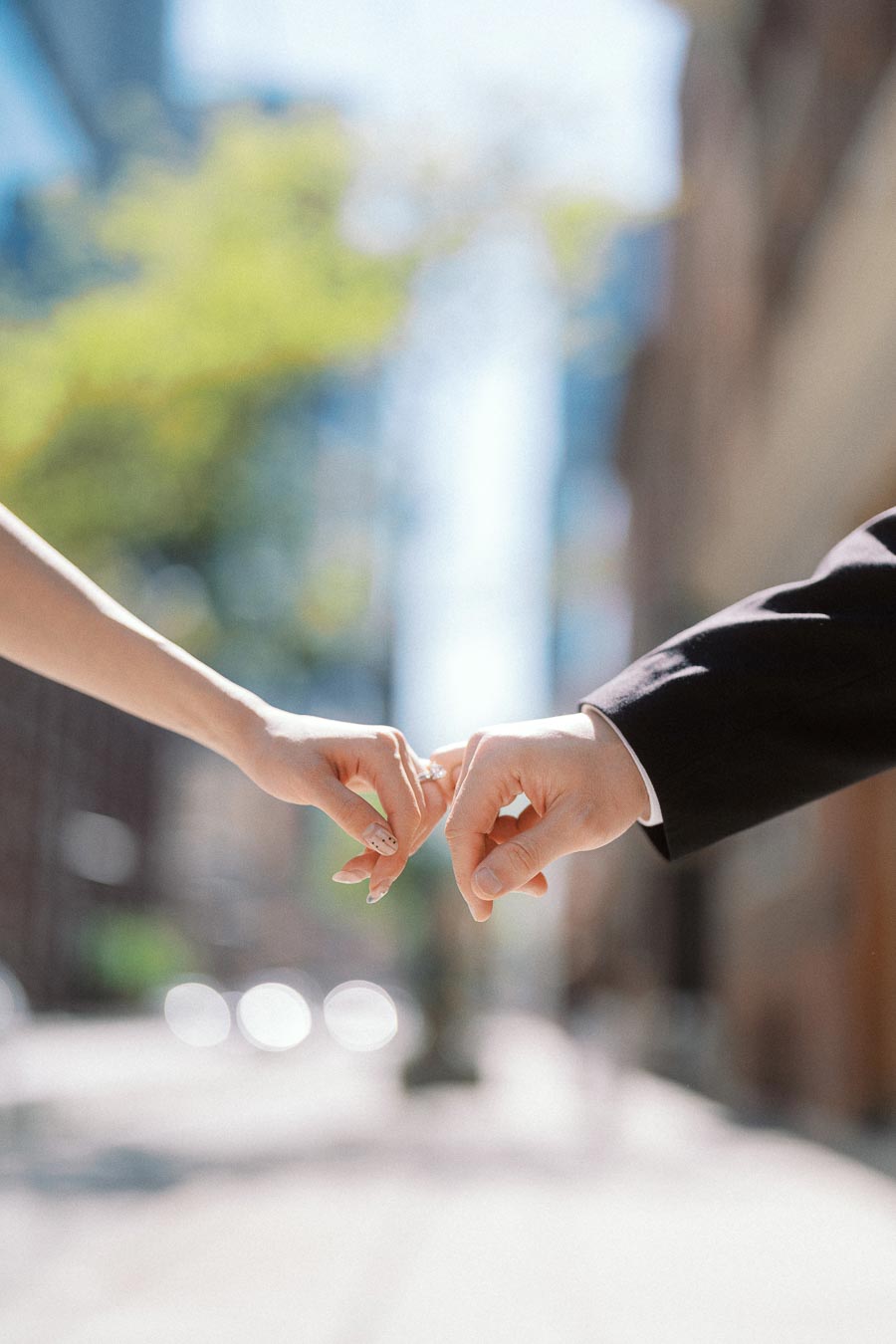 Two hands, one in a white sleeve and one in a black suit, gently touching fingers on a sunlit downtown street.