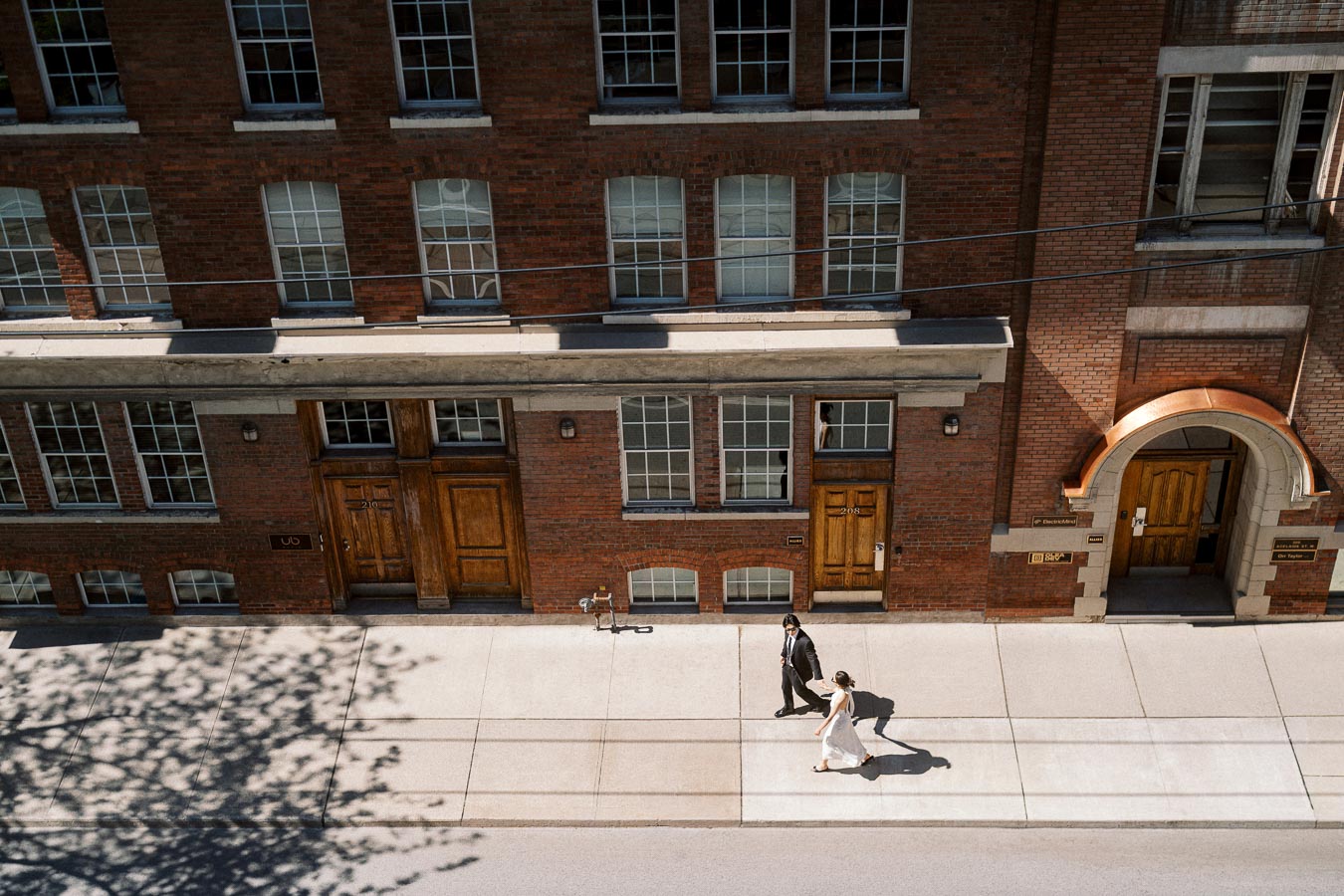 Couple walking together on a sunny sidewalk in front of a historic red brick building, photographed from above.