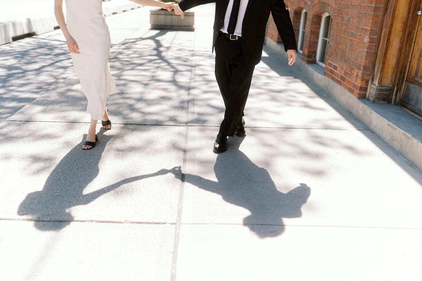 Two people walking hand in hand, casting long shadows on a bright sidewalk next to a brick wall.
