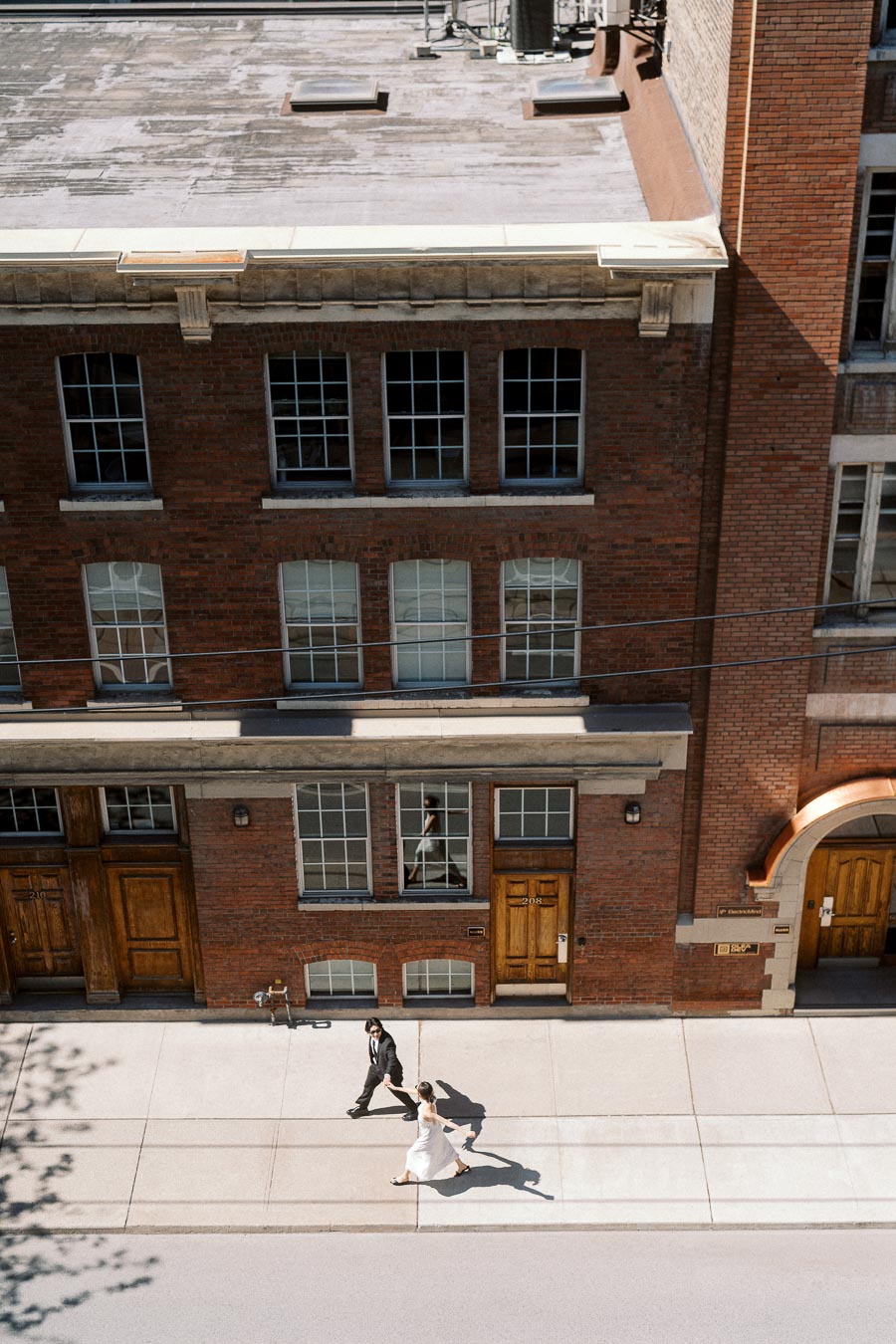 Couple holding hands and walking on a sunny sidewalk in front of a red brick building in downtown Toronto.