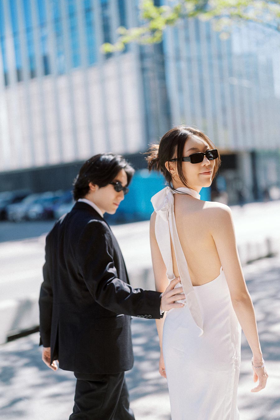 Couple wearing sunglasses walking hand in hand on a sunny downtown Toronto street with modern glass buildings in the background.