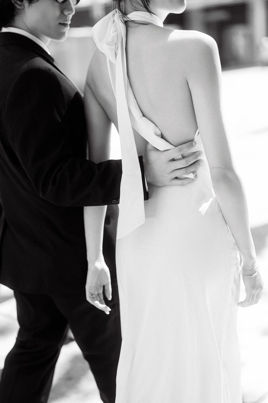 Close-up of a couple walking together, one wearing a white halter dress and the other in a black suit, holding each other affectionately.