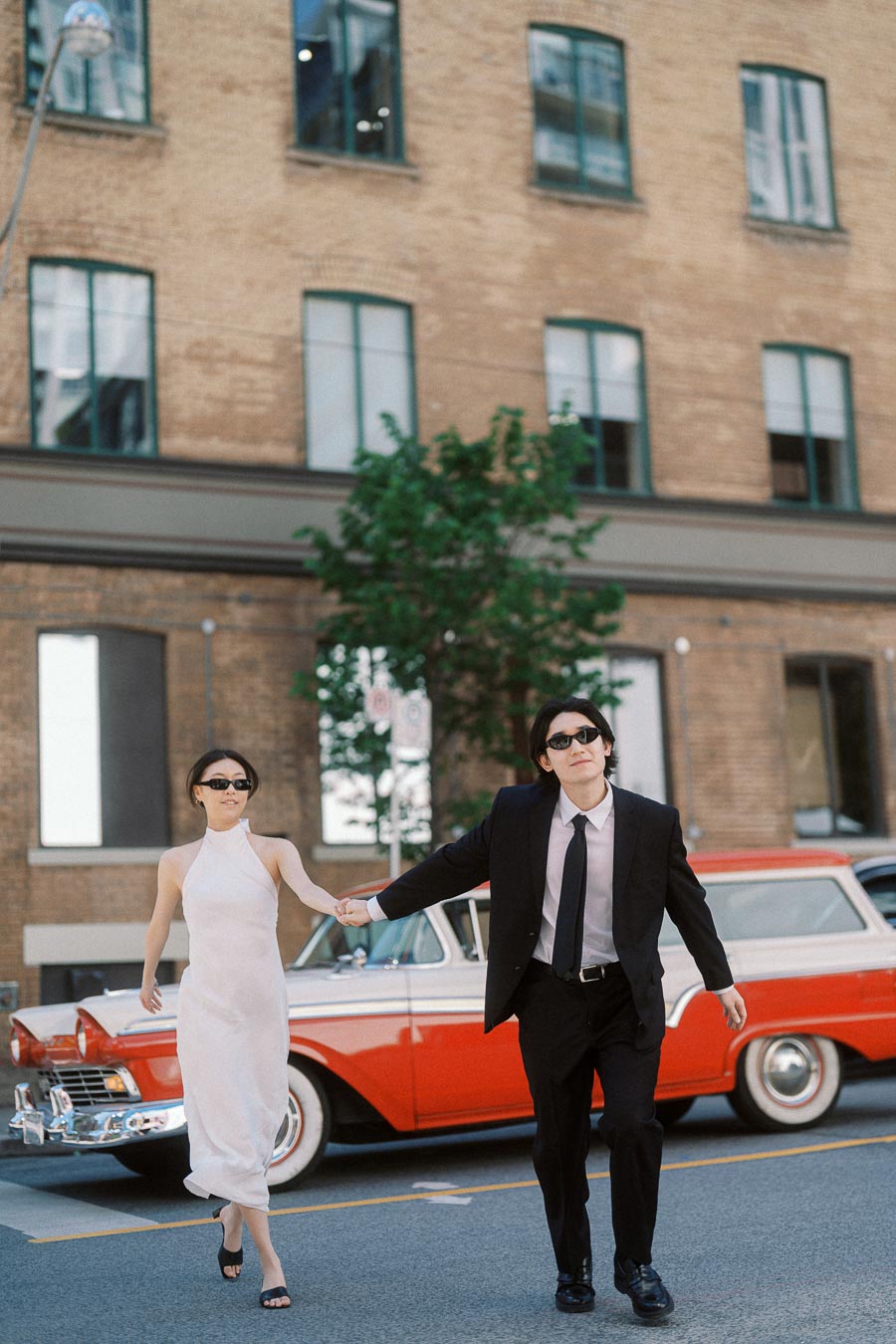 Couple in formal attire holding hands while crossing a city street, vintage red car and brick building in background.