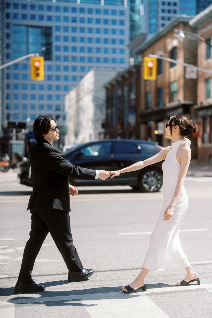A couple, one in a black suit and one in a white dress, holding hands while crossing a downtown Toronto street with modern buildings in the background.