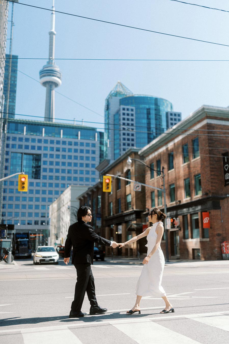 Couple holding hands and walking across a downtown Toronto crosswalk with the CN Tower and city buildings behind them.