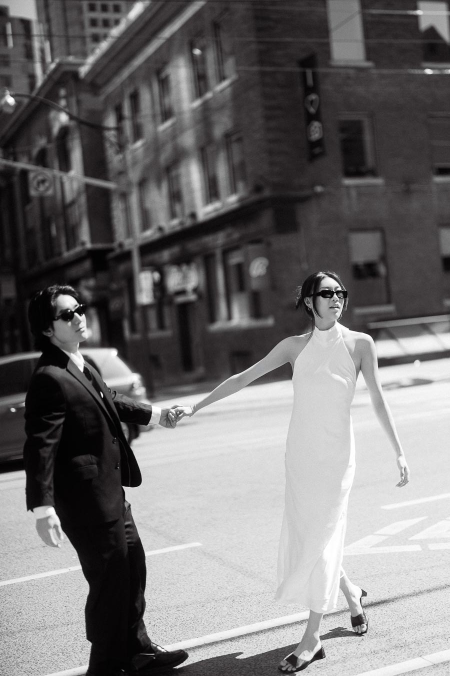 A stylish couple holding hands and walking across a city street, both wearing sunglasses, with tall brick buildings in the background.