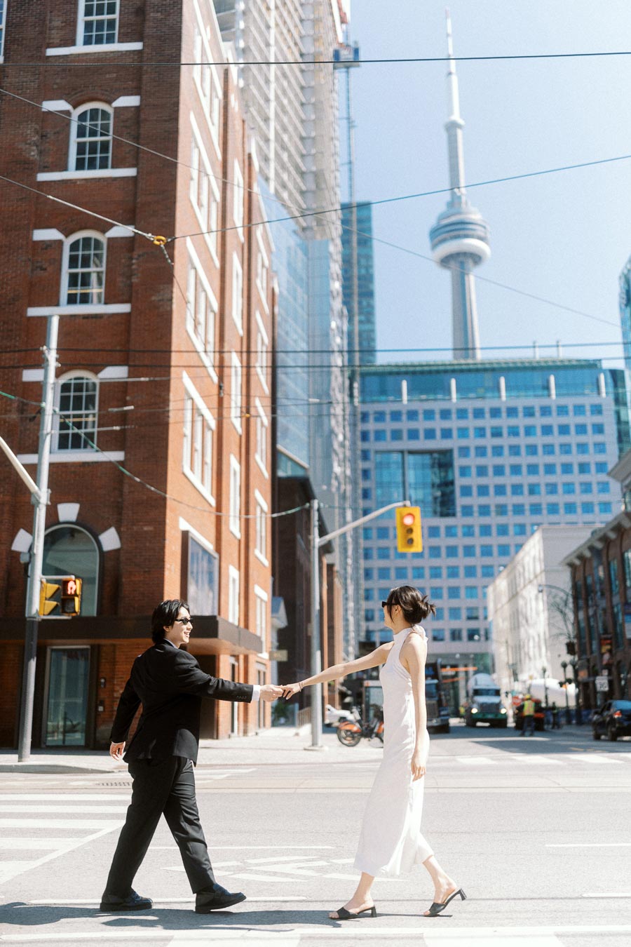 Couple holding hands and crossing a city street in downtown Toronto, with the CN Tower and urban buildings visible behind them.