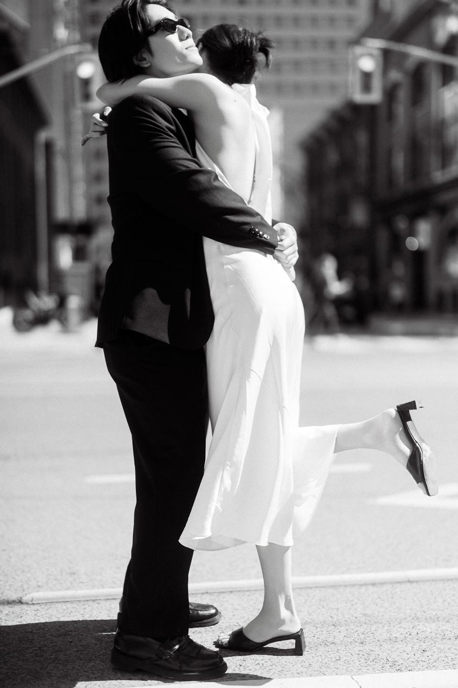 Couple embracing in the middle of a city street, one person in a white dress lifting their foot, both wearing sunglasses, with tall buildings in the background.