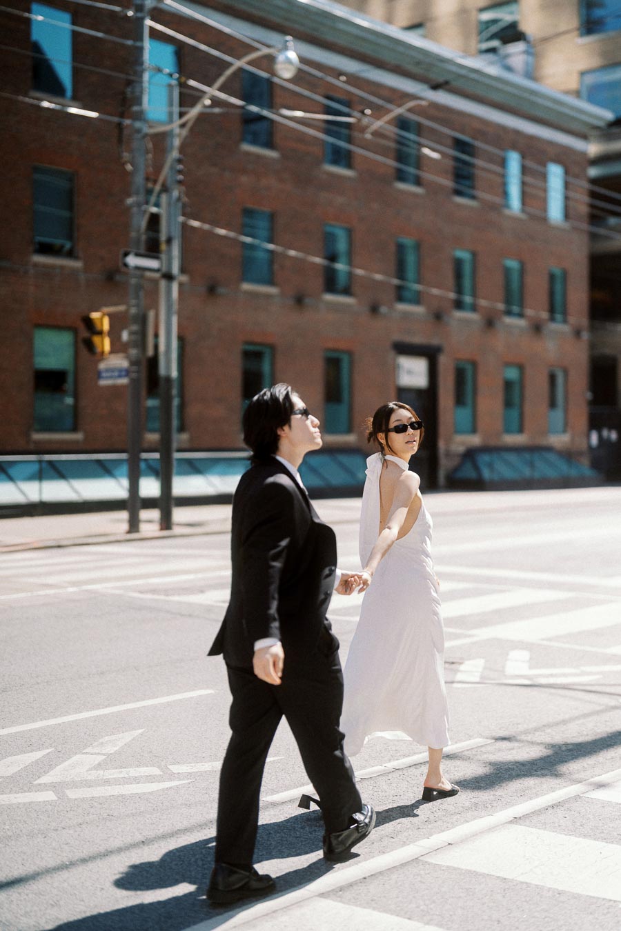 Couple holding hands and crossing a city street in Toronto, wearing formal outfits and sunglasses, with a brick building in the background.