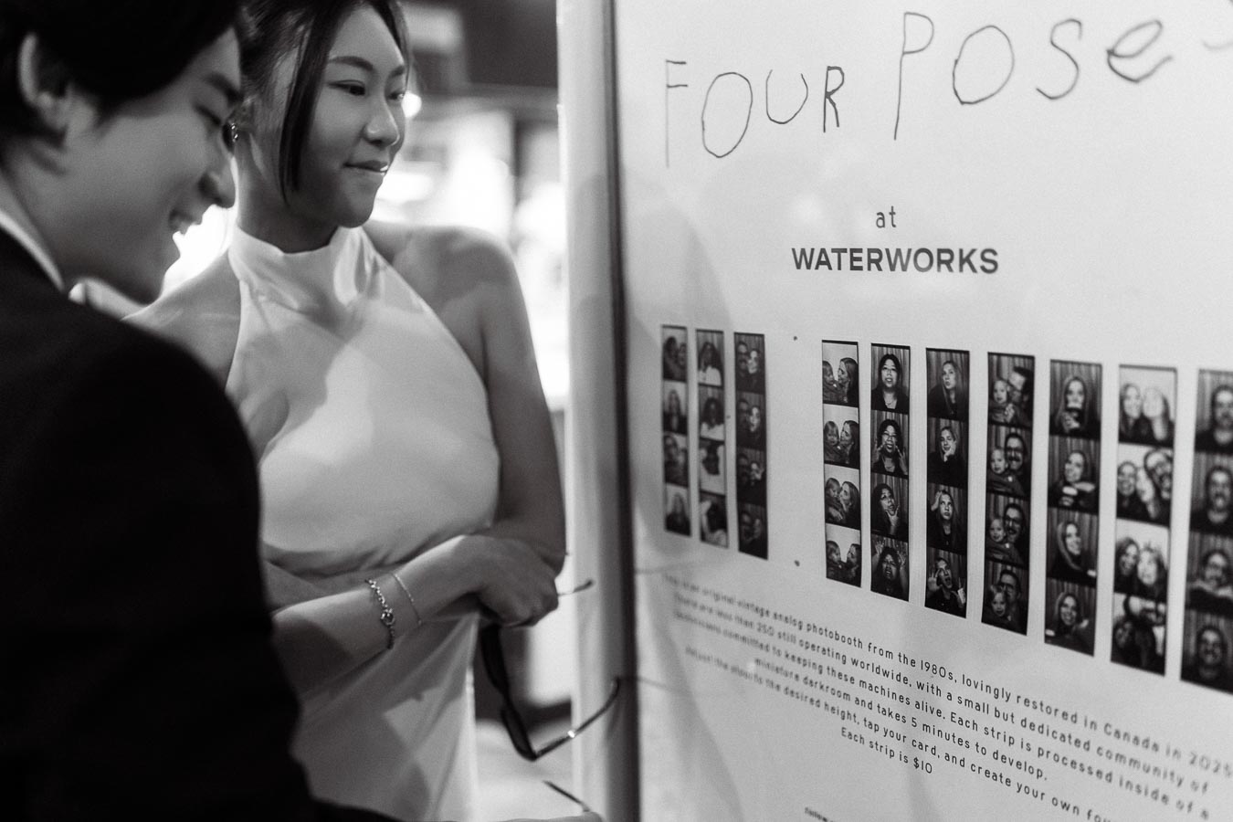 Jun and Alex stand together, smiling and looking at a wall of photo booth portrait strips in a candid moment.