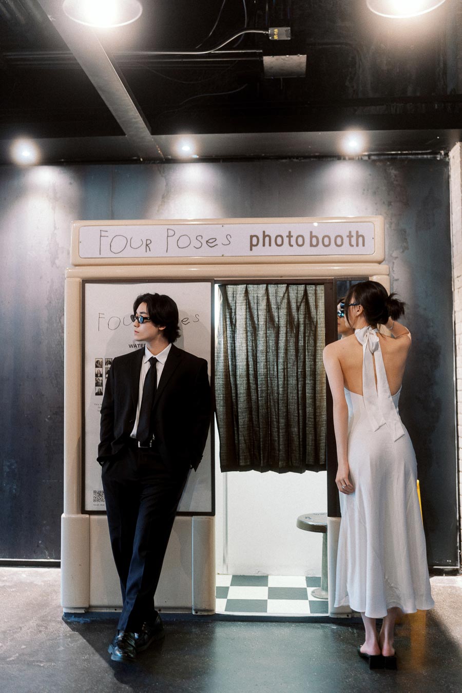 Couple in formal attire posing stylishly by a vintage photobooth, with moody lighting and a city editorial vibe.