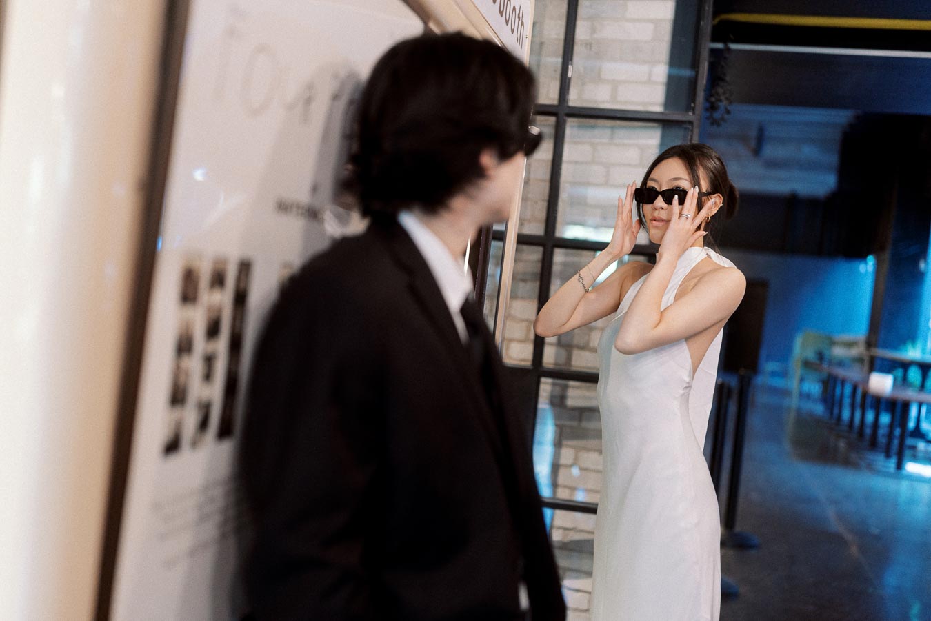 Woman in a white dress adjusting sunglasses while man in a black suit looks on by a vintage photo booth in a modern indoor setting.