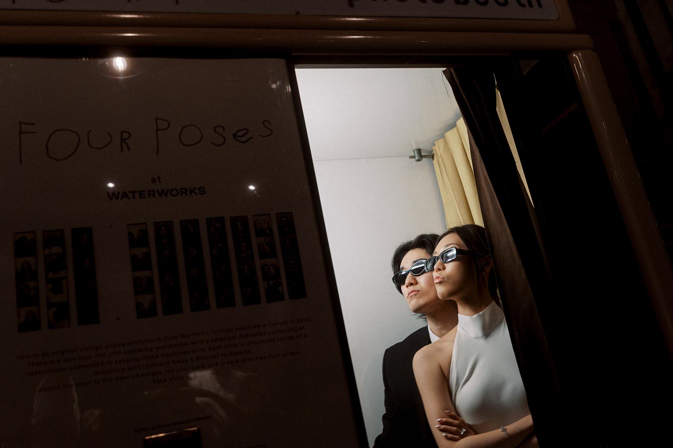Couple wearing sunglasses posing together in a vintage photo booth, with stylish outfits and dramatic lighting.