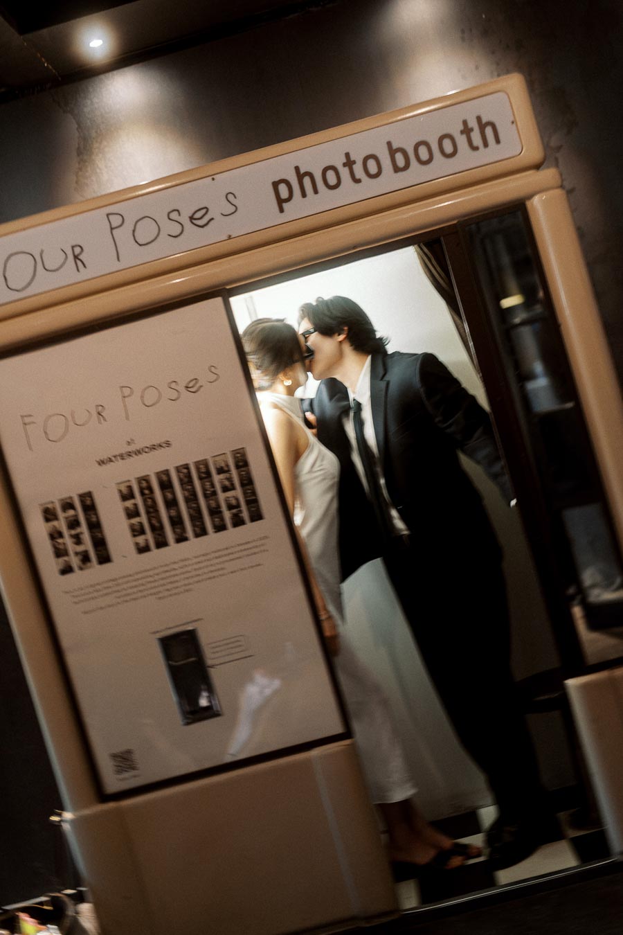 A couple in formal attire kisses inside a vintage photobooth, illuminated by soft light, creating a candid and romantic scene.