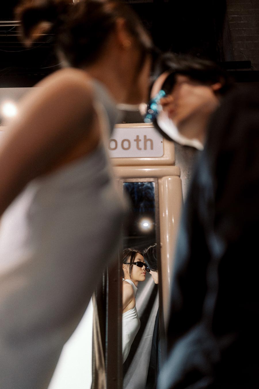 A couple in sunglasses leans toward each other, reflected in a vintage photo booth mirror in a moody, urban setting.