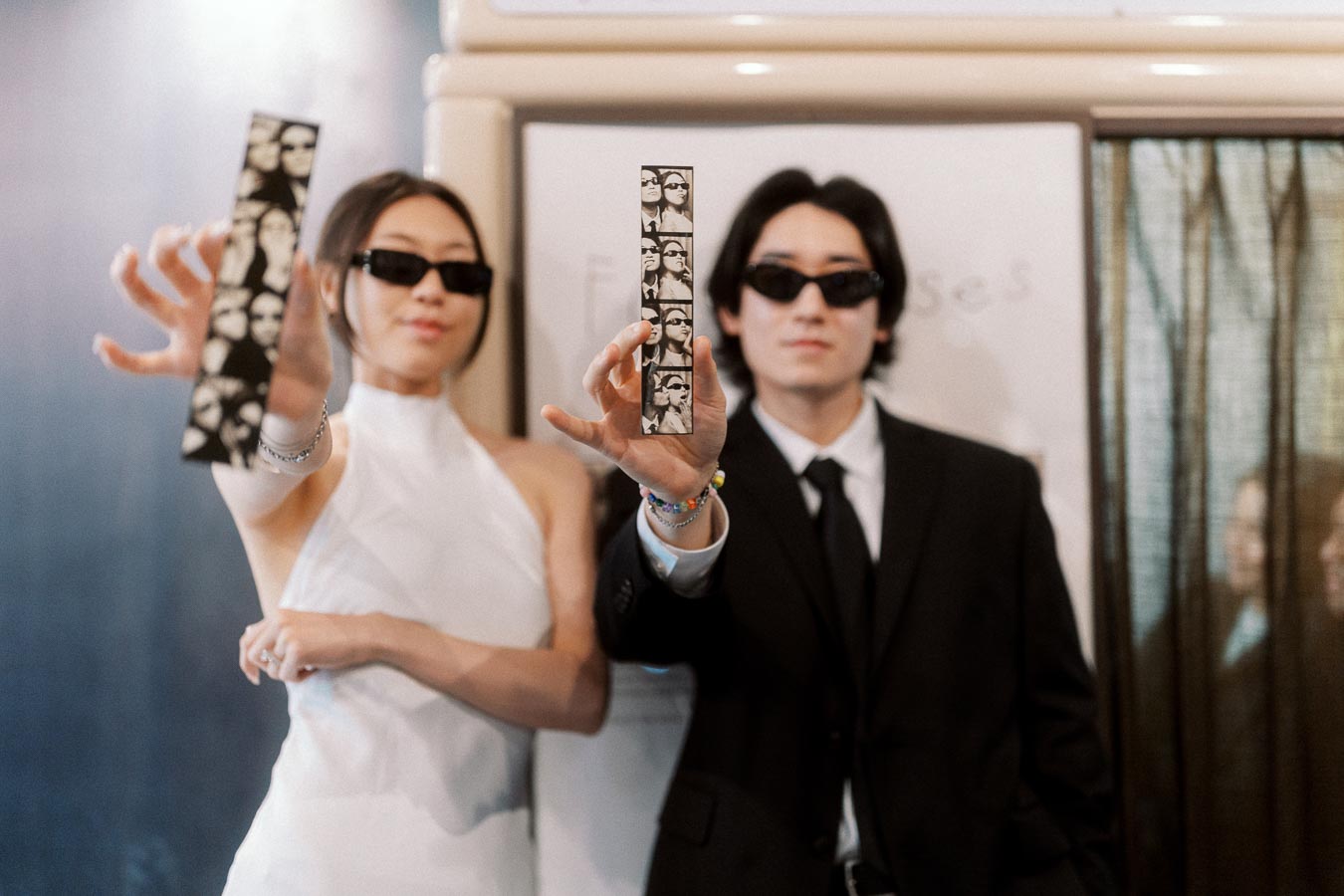 Couple in formal attire and sunglasses holding photo booth strips in front of a vintage booth, smiling together.
