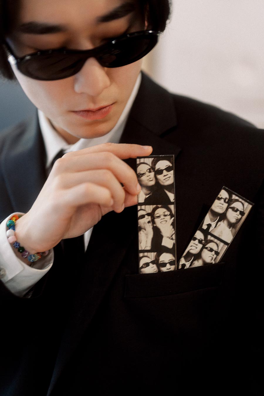 Person in black suit and sunglasses holding photo booth strips with couple portraits in suit pocket.