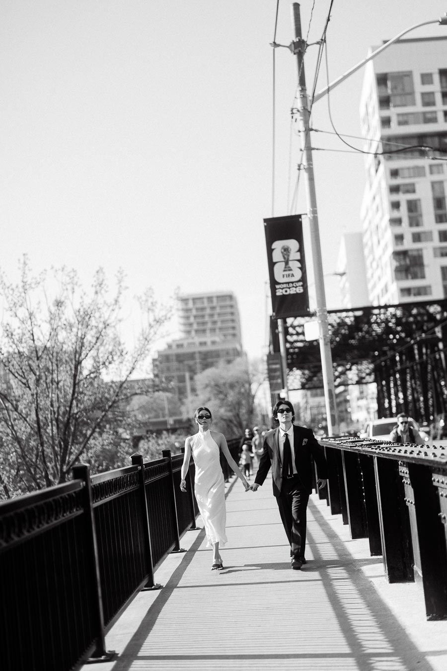 Couple wearing sunglasses, holding hands while walking on a city bridge with modern buildings in the background.