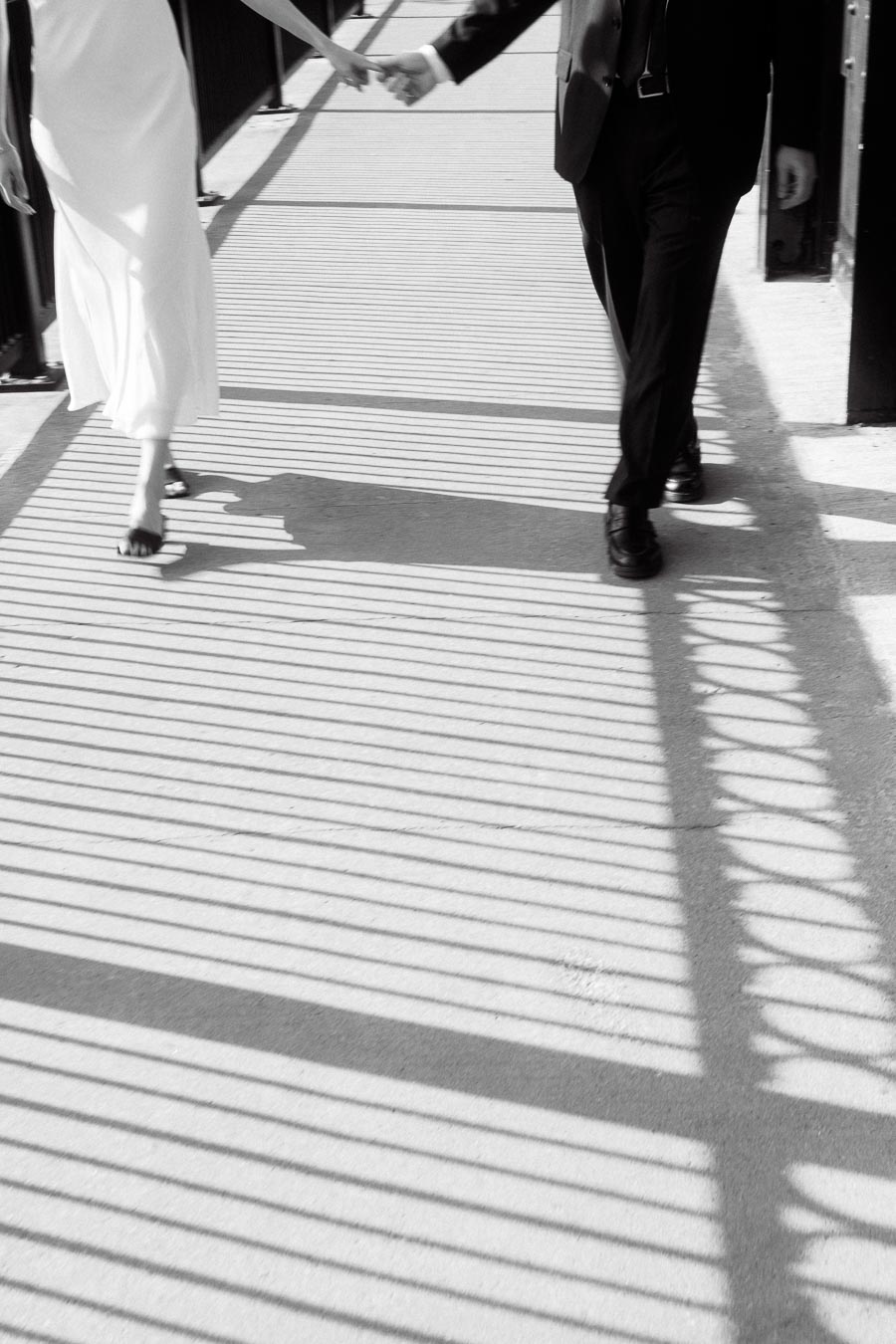 Two people holding hands walking on a bridge with dramatic striped shadows on the ground, wearing a white dress and a black suit.