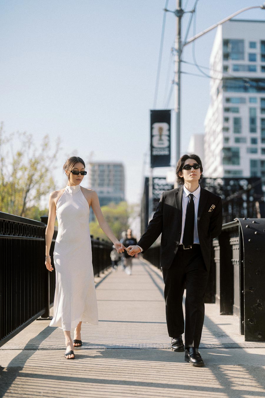 Couple holding hands walking on Sir Isaac Brock Bridge in downtown Toronto, both wearing sunglasses and formal outfits, with urban buildings in the background.