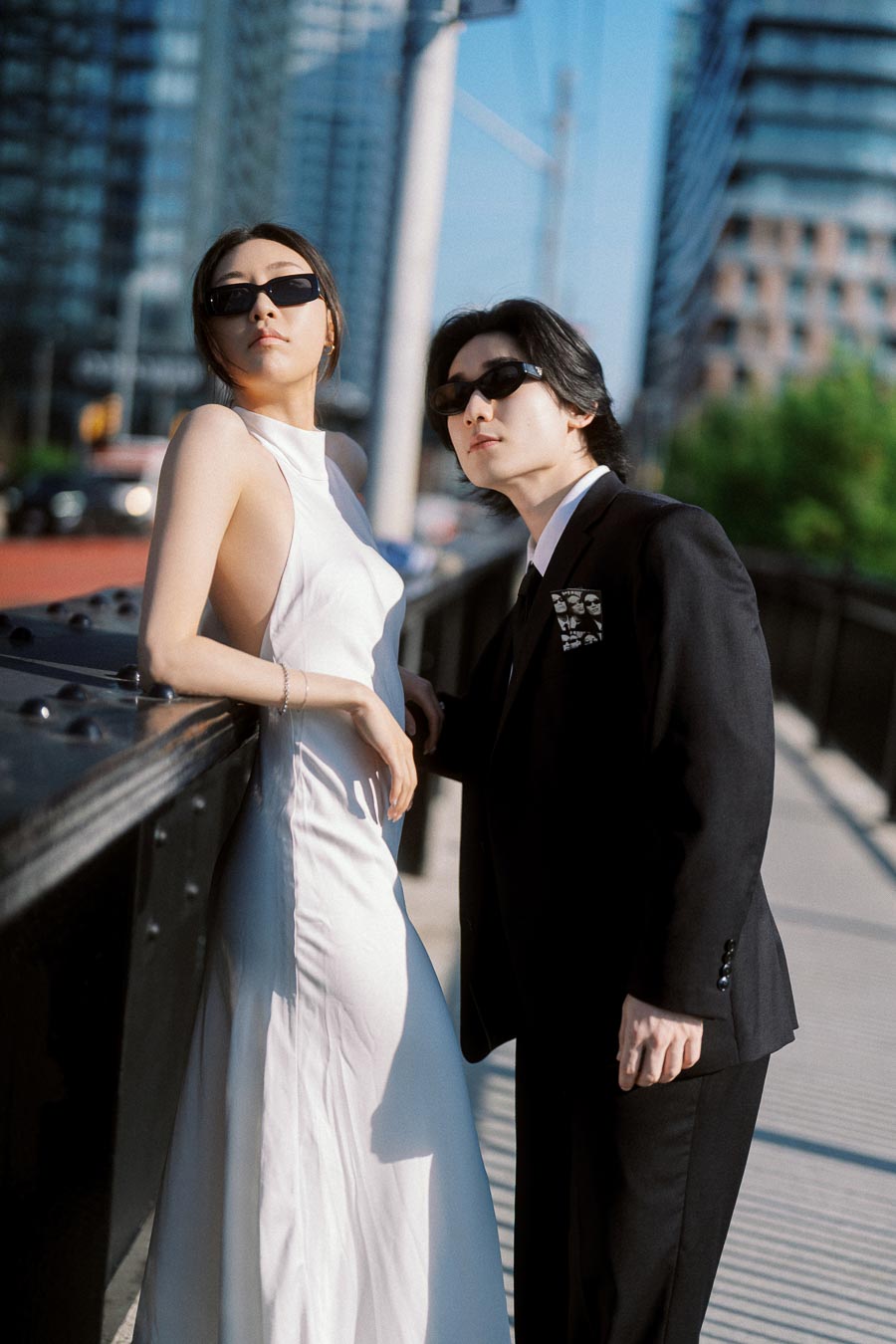A stylish couple wearing sunglasses, one in a white dress and the other in a black suit, pose together on a downtown Toronto bridge with modern buildings in the background.